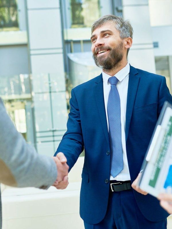 Portrait of bearded middle-aged entrepreneur with toothy smile shaking hand of his business partner after successful completion of negotiations, interior of modern office lobby on background
